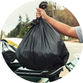 Hand holding a full black trash bag above a green bin, outdoors.
