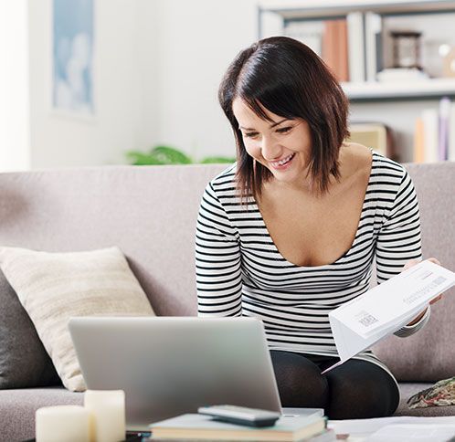 Woman on couch using laptop, holding papers, smiling.