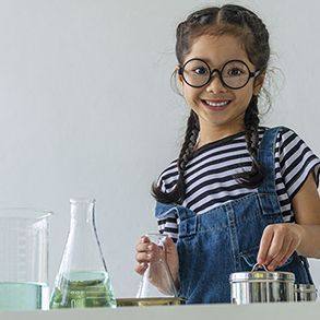 Girl with glasses smiles while conducting a science experiment with beakers and a metal container.