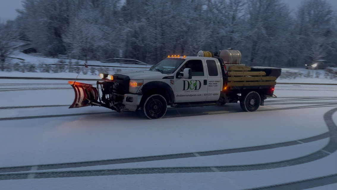 White snow plow truck plowing a snowy parking lot.