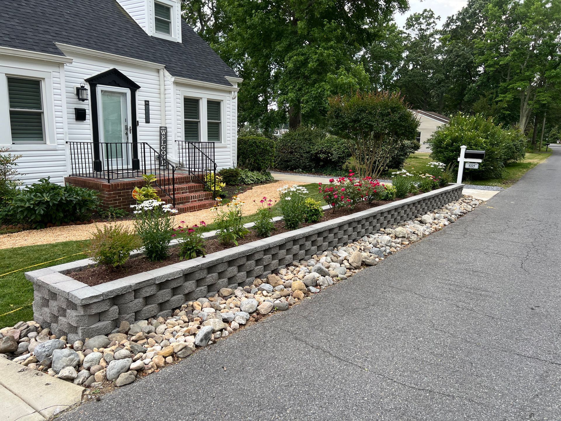 House with flower bed alongside a street; gray retaining wall with flowers and rocks.