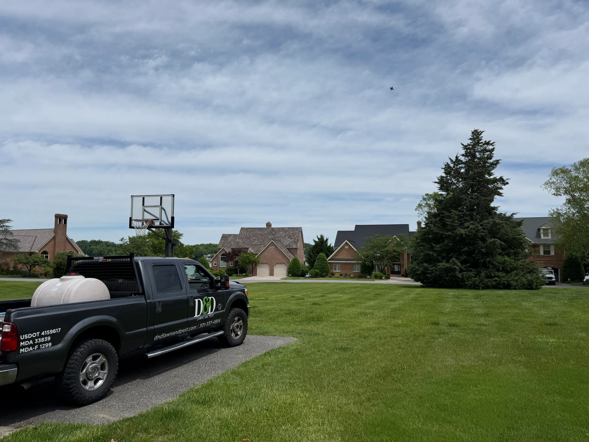 Dark truck with a white tank parked on a gravel driveway in front of a green lawn and houses under a cloudy sky.