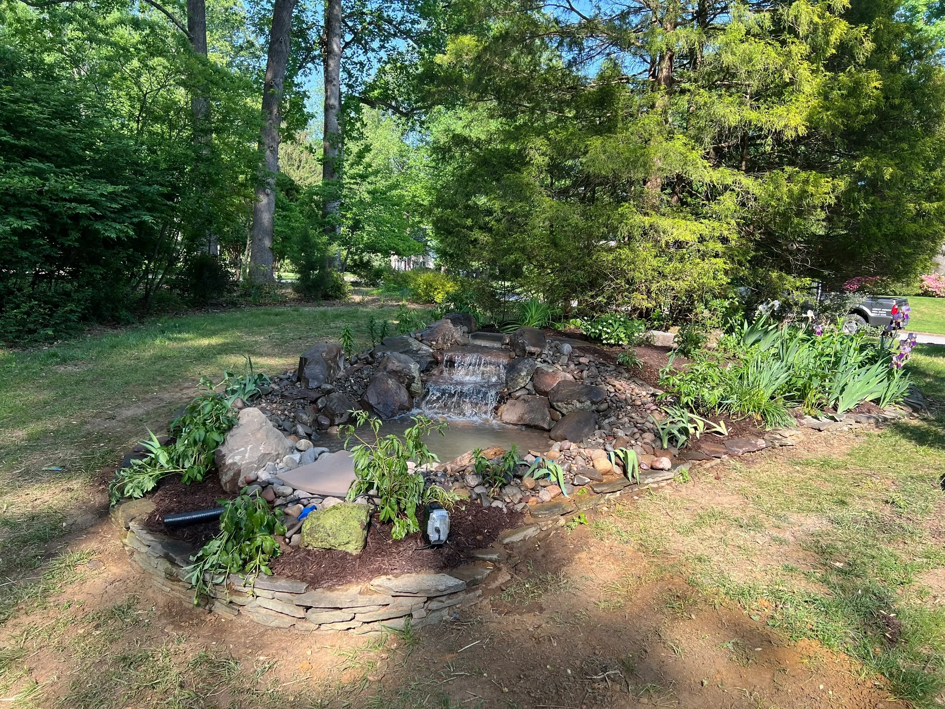 Small backyard waterfall feature surrounded by rocks, plants, and greenery.