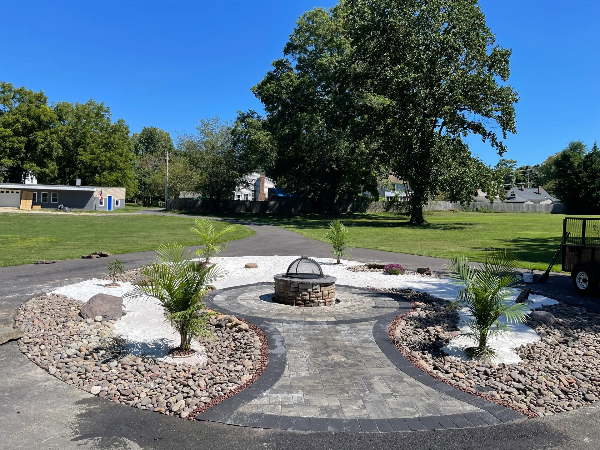 Fire pit surrounded by paving stones, rocks, and white stone. Green trees and clear sky in the background.