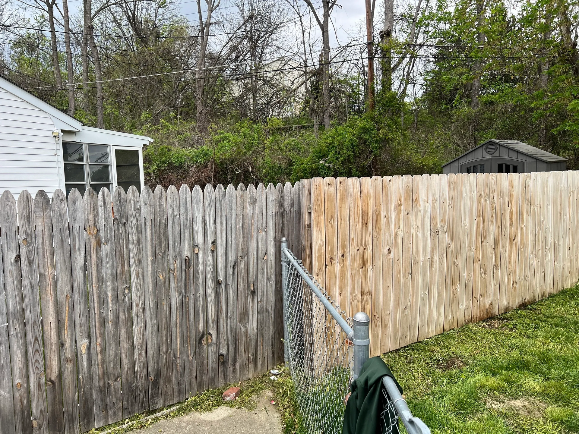 Two wooden fences with a chain-link gap. One weathered, one new. A small shed and trees in the background.