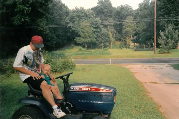 Man wearing red cap, sits with toddler on riding lawnmower in front yard. Green grass and trees visible.