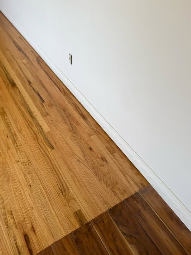 Hardwood floor meeting a white wall with a visible electrical outlet and baseboard trim