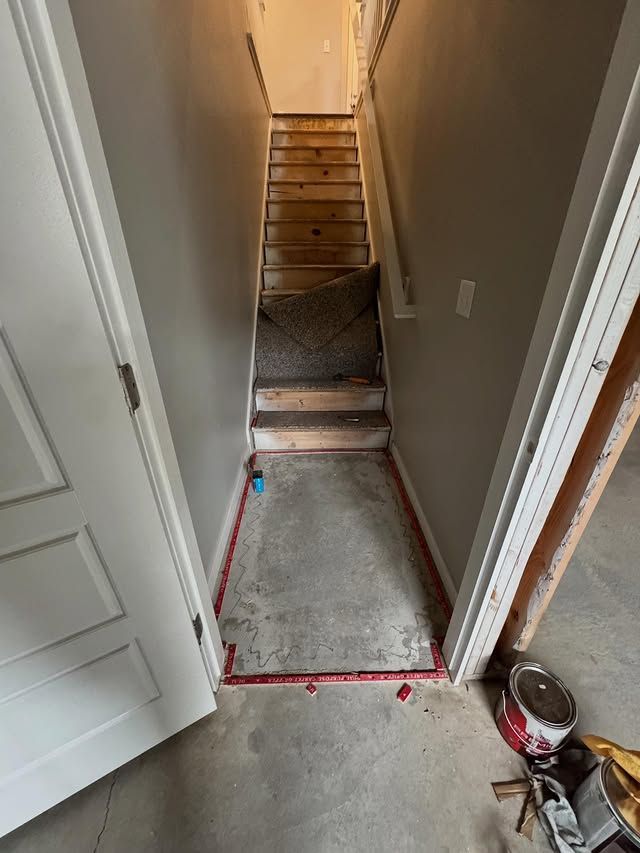 Narrow hallway with red-taped floor markings leading to basement stairs, showing a carpeted stairway and doorway.