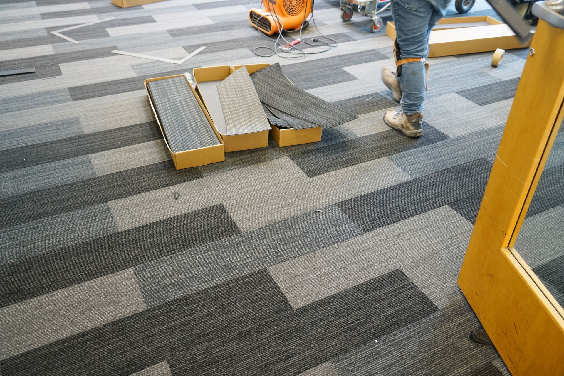 Carpet tiles being installed in a hallway, with a worker’s legs and tools visible on the floor