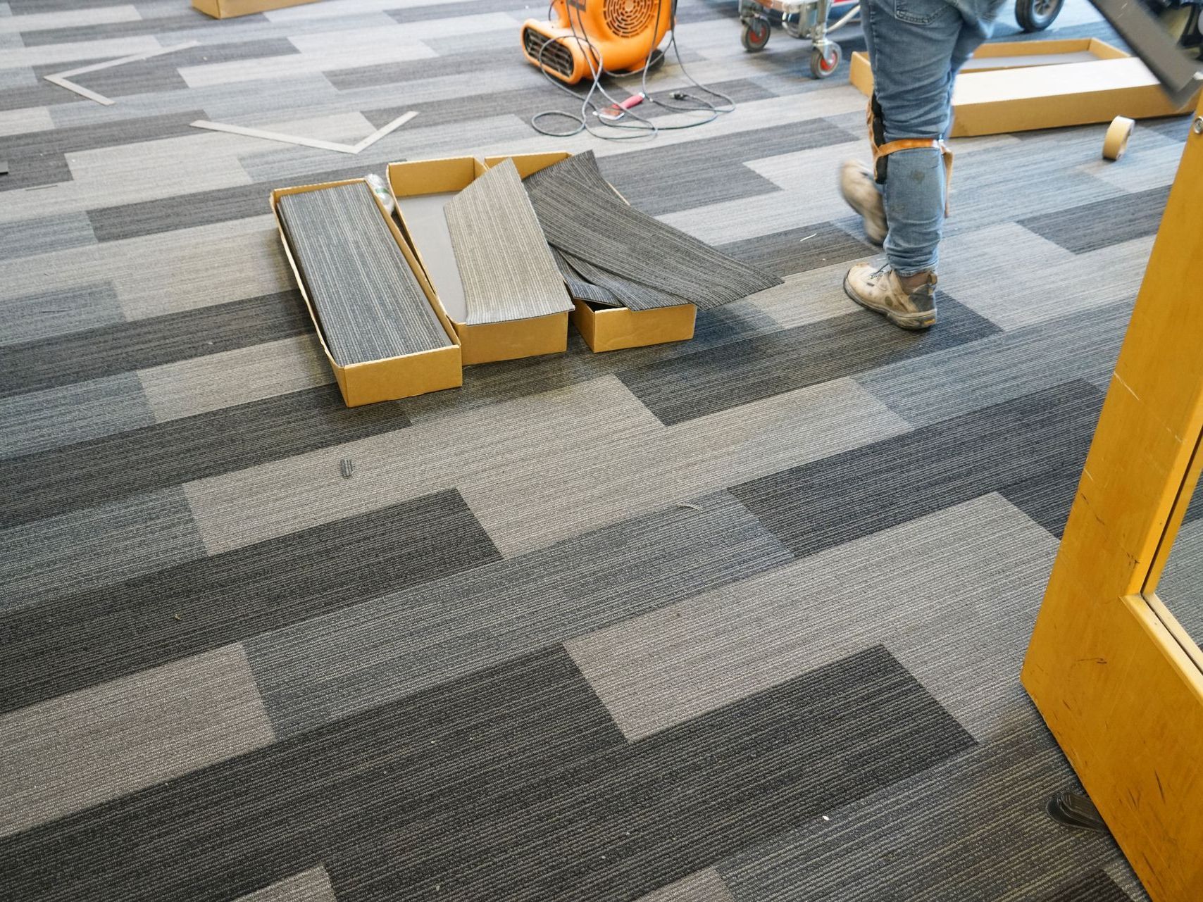 Worksite floor with a folded yellow step ladder and people’s legs on gray patterned carpet