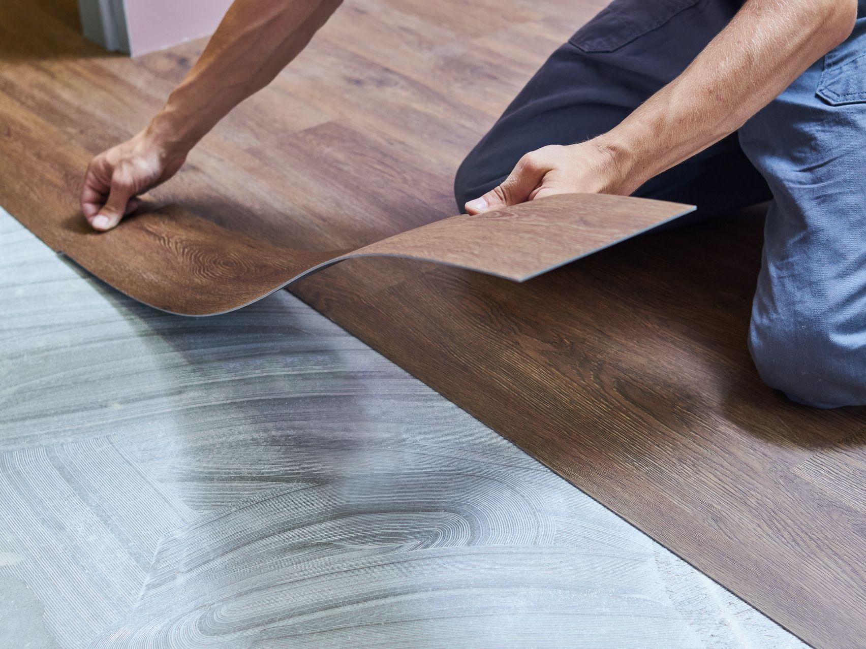 Hands laying dark wood-look flooring over a light subfloor area