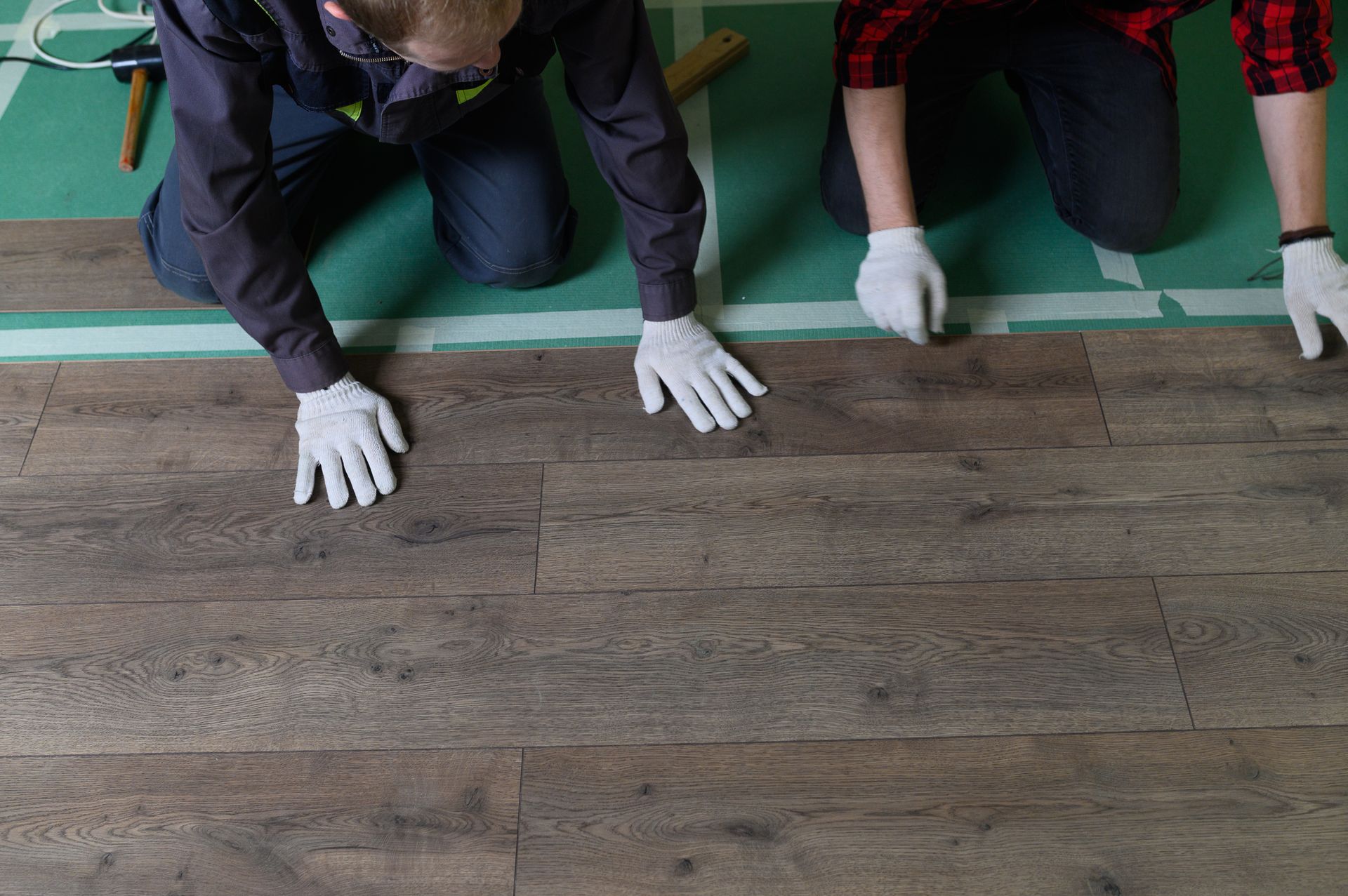 People kneeling on a green mat, wearing gloves, working on a wood-look floor surface