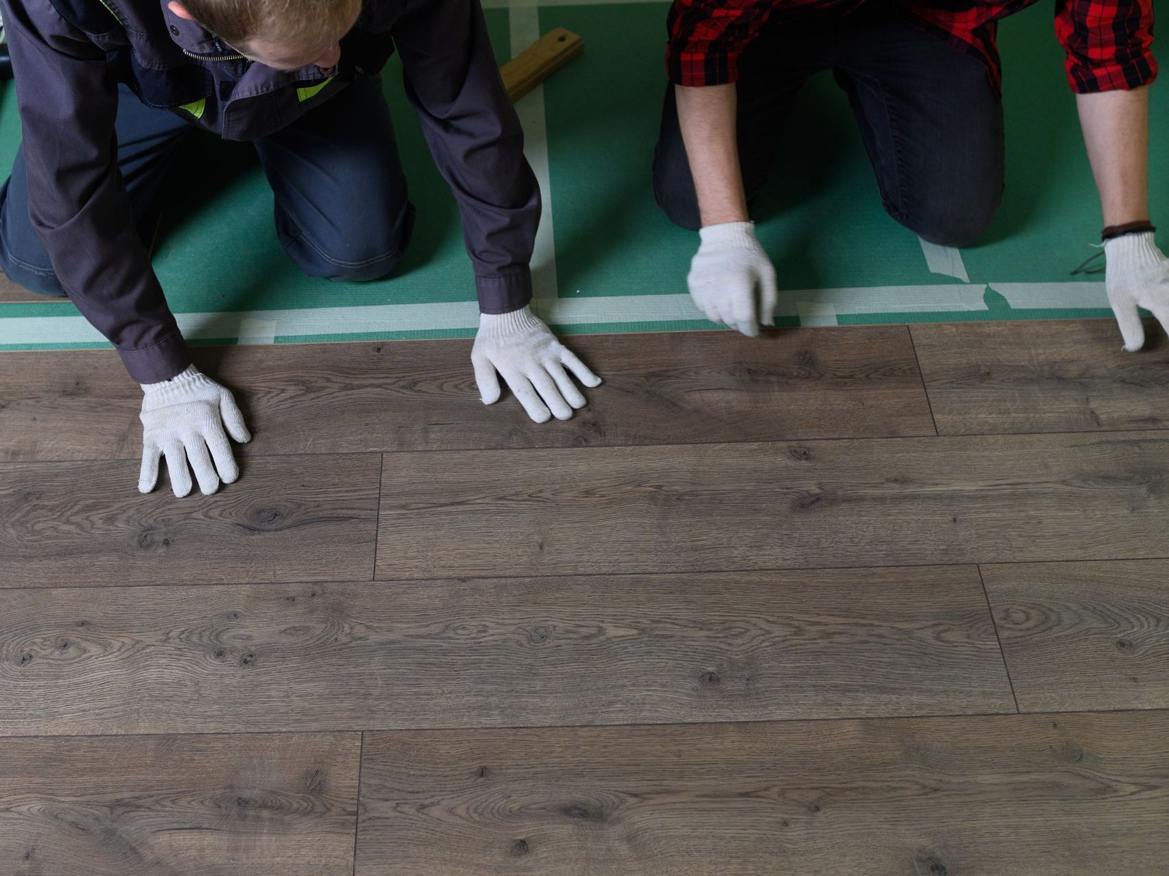 Three people kneel on a floor, wearing white gloves and working on a wood-patterned surface.