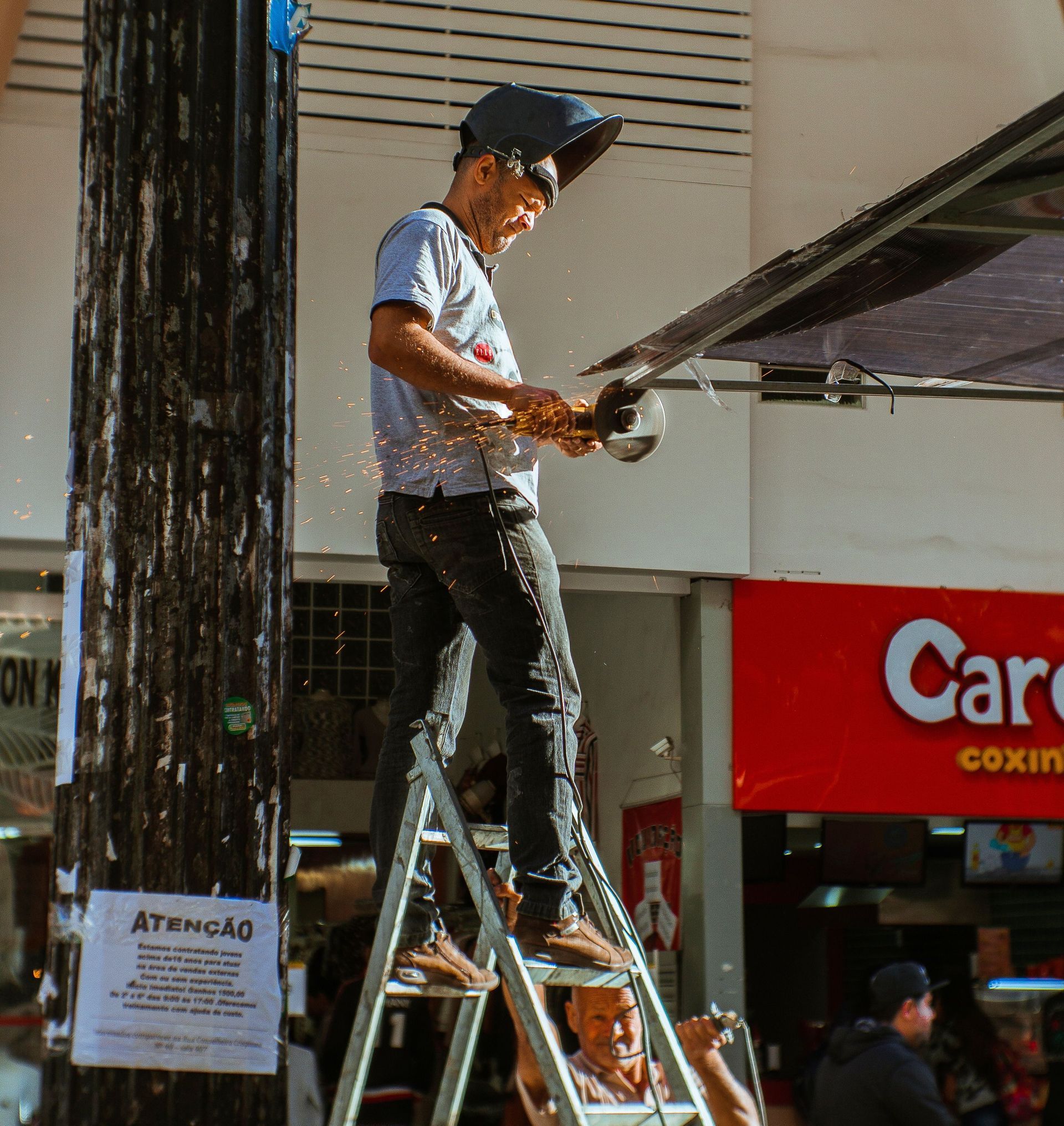 Worker on a ladder using a power tool to install or adjust a large outdoor sign on a city street.