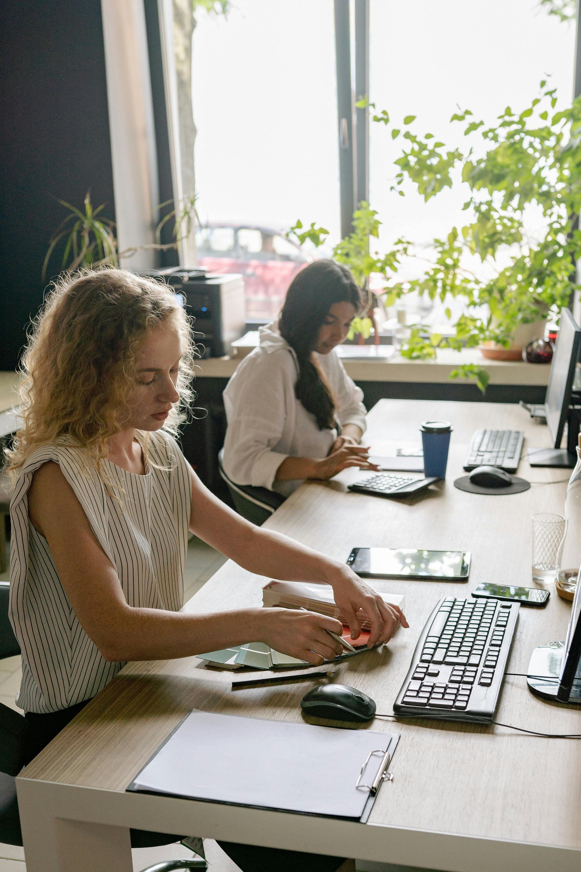 Two people working at desks in a bright office, using a keyboard and laptop near a window.