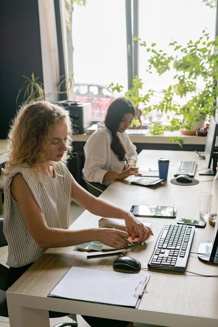 Two people working at desks in a bright office, using a keyboard and laptop near a window.
