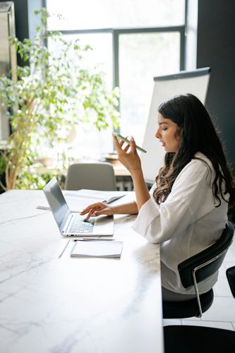 Woman working at a desk with laptop and phone in a bright office, near a window and flip chart