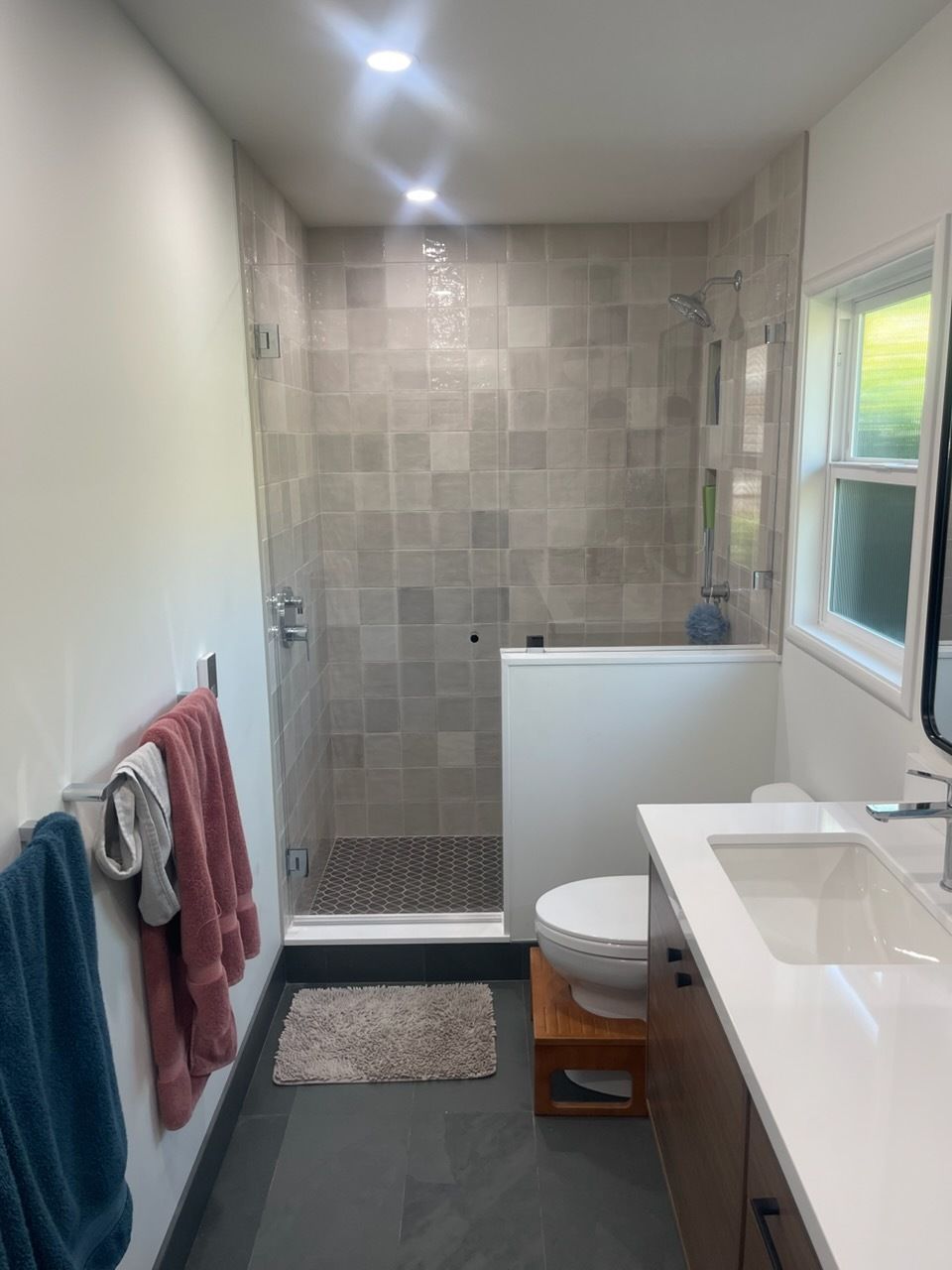 Narrow bathroom with walk-in shower, toilet, vanity, window, and towels on wall racks.