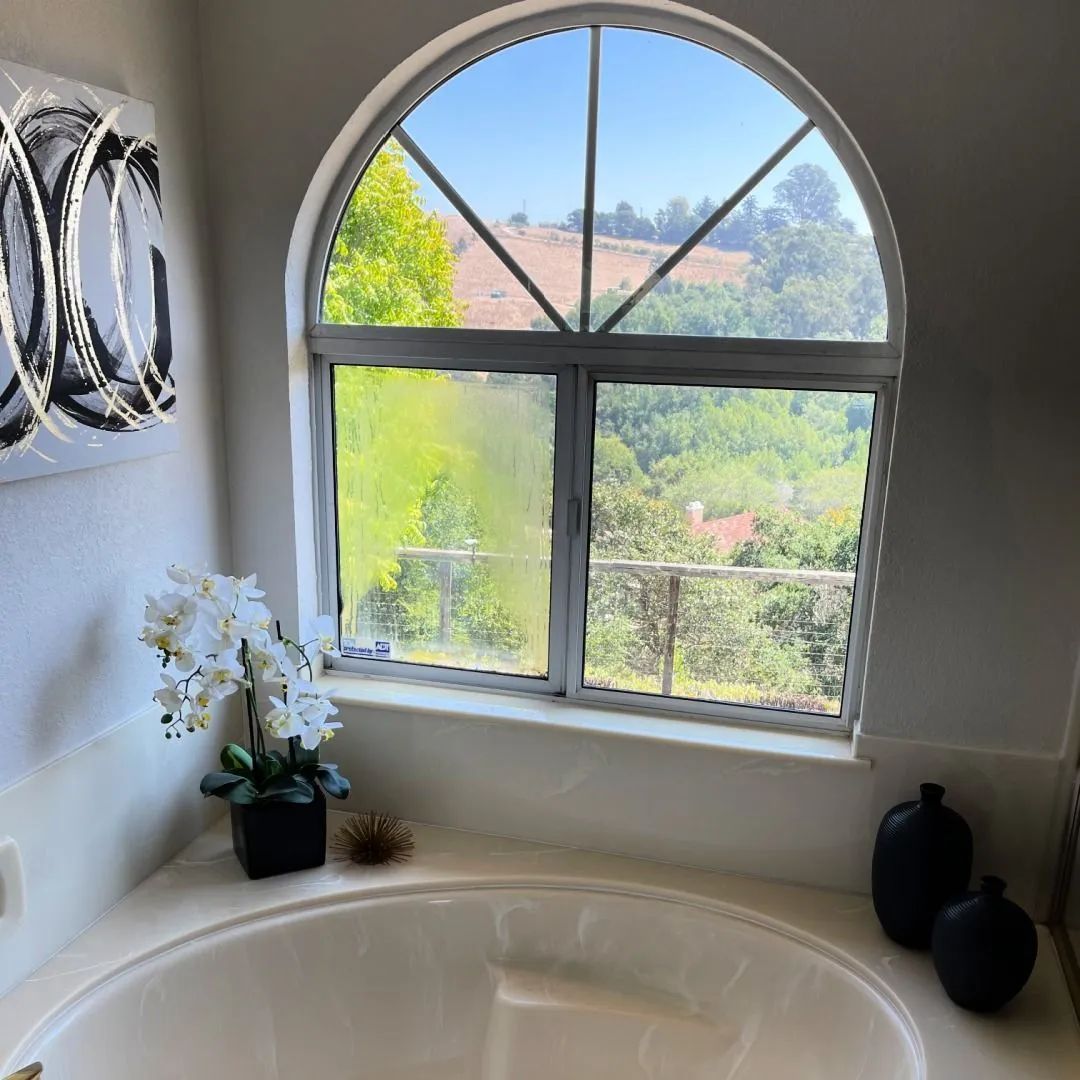 Bathroom with a rounded window overlooking a green landscape, white tub, and decor.