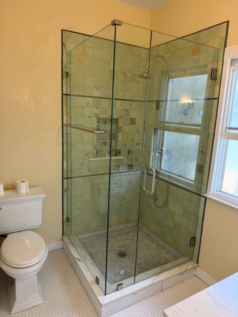 Corner glass shower enclosure in a bathroom beside a toilet and window, with beige tile walls.
