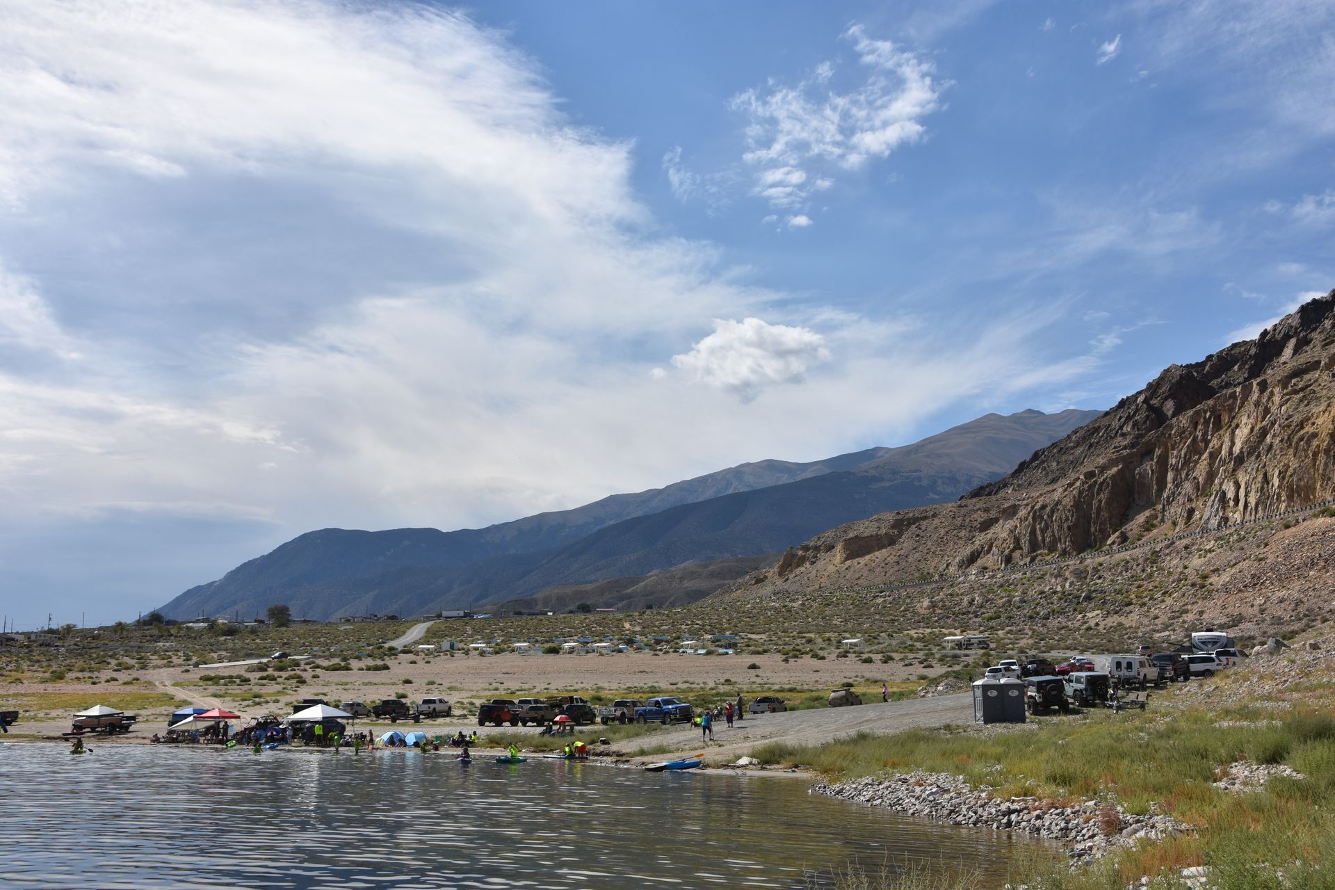 A lake with mountains in the background and a lot of cars parked on the shore.