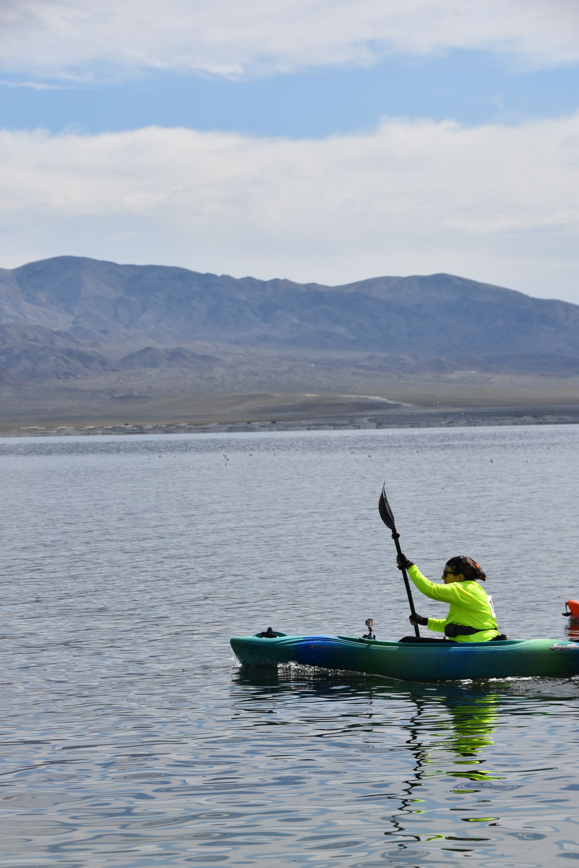 A person is paddling a kayak on a lake with mountains in the background.