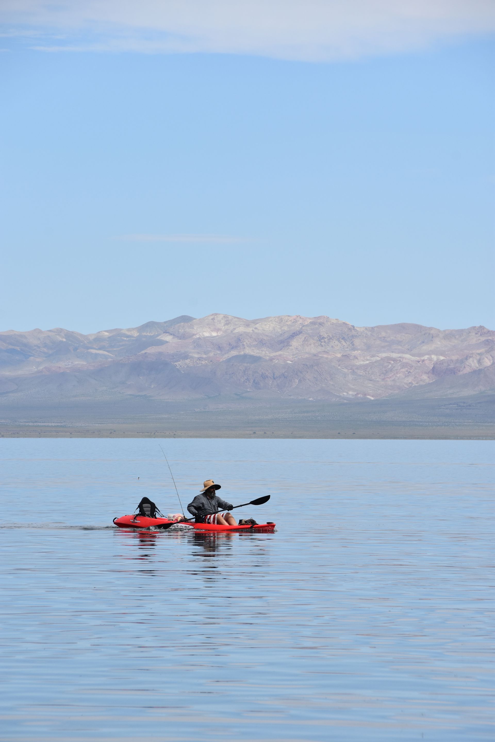 A man is paddling a red kayak on a lake with mountains in the background.