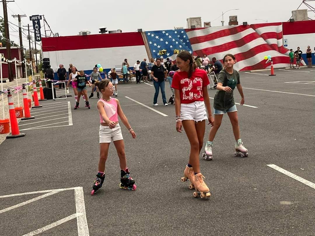 Three girls roller skating in the Nevada Joe's parking lot with it's American flag mural in the background in Hawthorne, NV