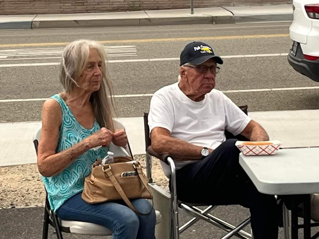 Elderly couple  watching Skate Night at Mineral Co, NV. The woman holds a purse, the man wears a hat and has a hot dog.