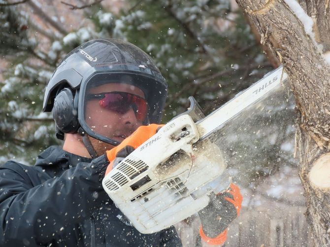Person in a helmet using a chainsaw to cut a tree branch in snowy conditions