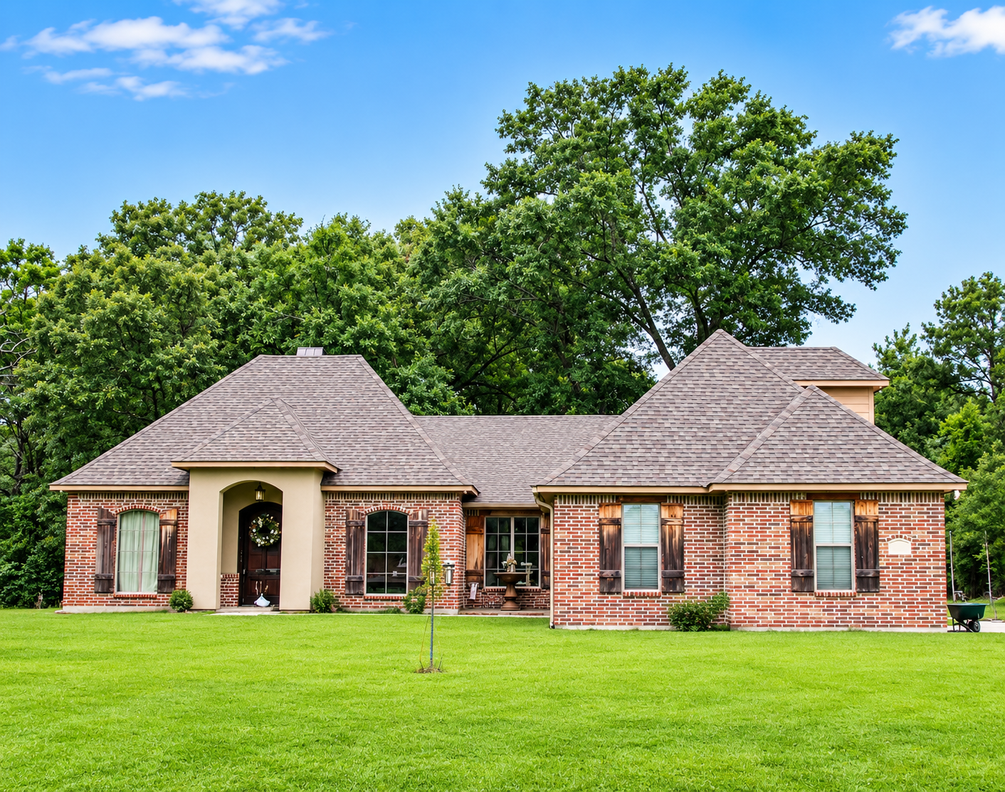 Brick suburban house with gray shingled roof, arched front entry, and large green lawn under blue sky