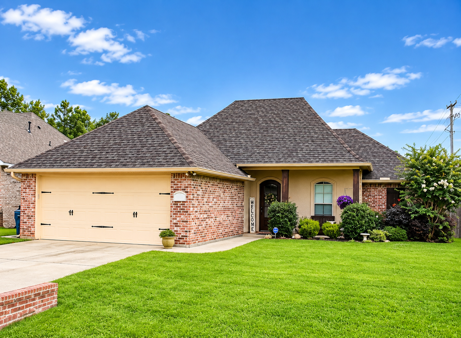 Single-story brick house with a beige garage, dark shingle roof, and green lawn under a blue sky