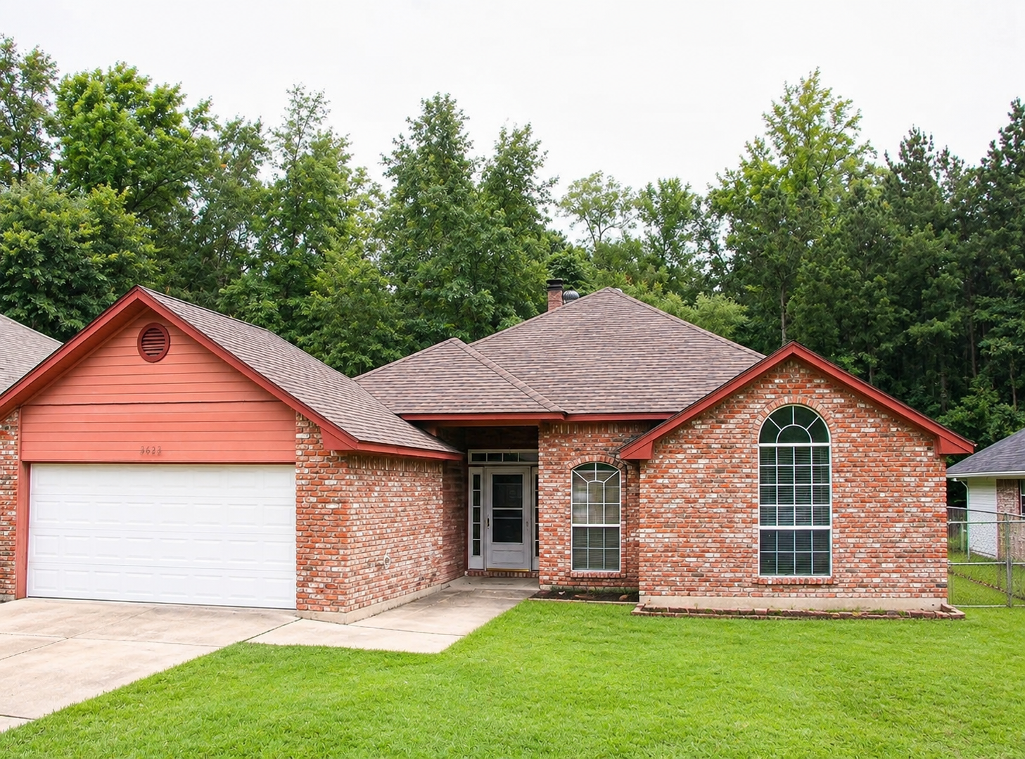 Brick suburban house with red trim, white garage door, and green lawn under trees