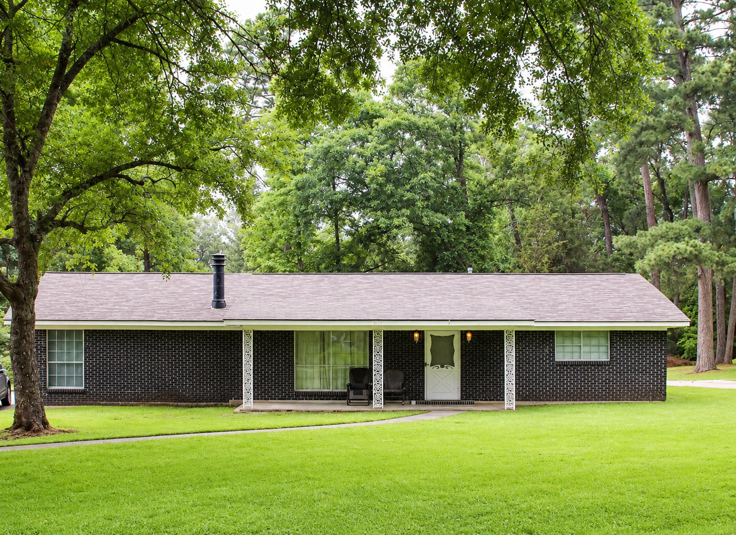 Single-story brick house with a gray roof, surrounded by large green trees and a grassy lawn.