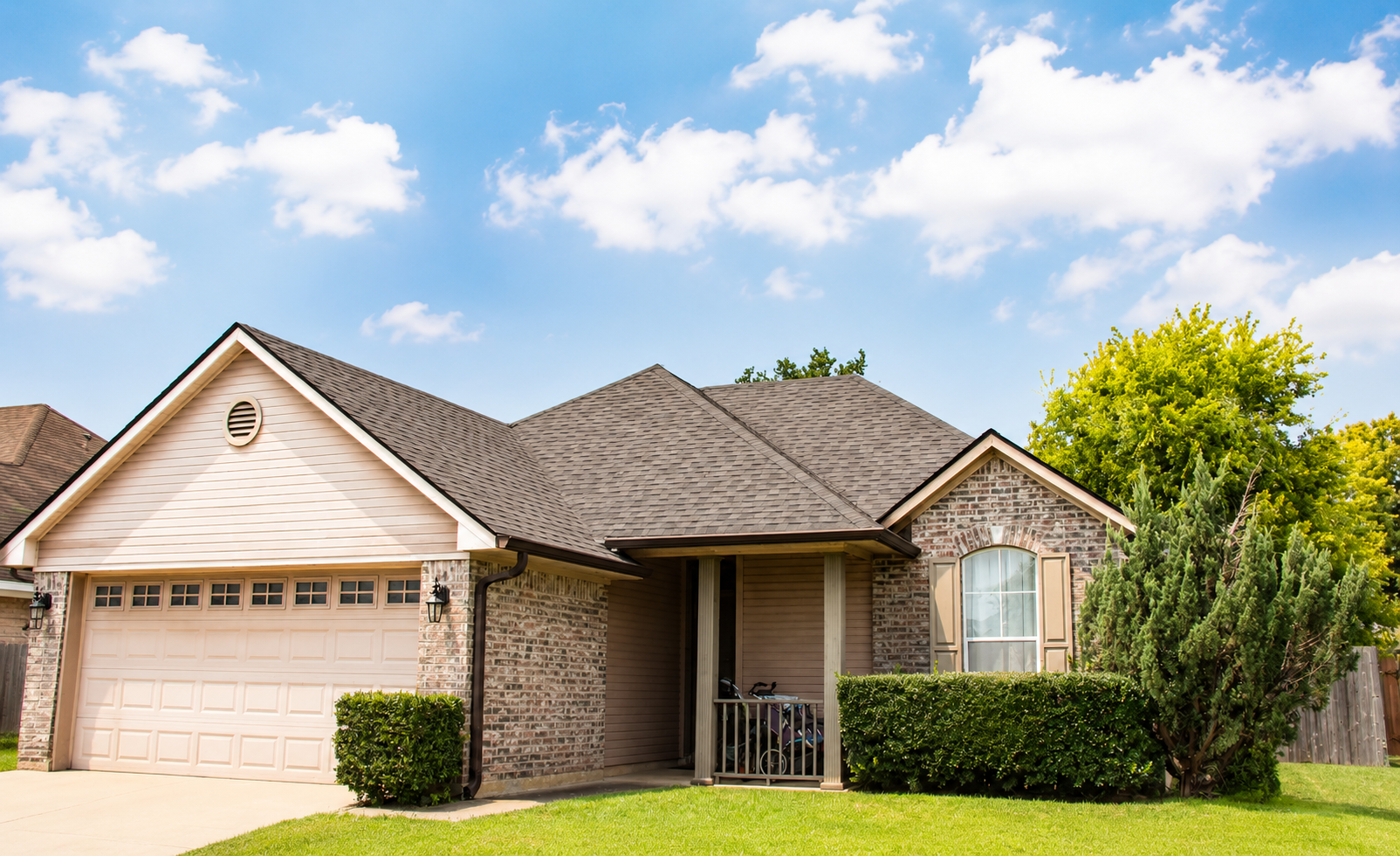 Single-story suburban brick house with beige siding, attached garage, and a green lawn under a blue sky