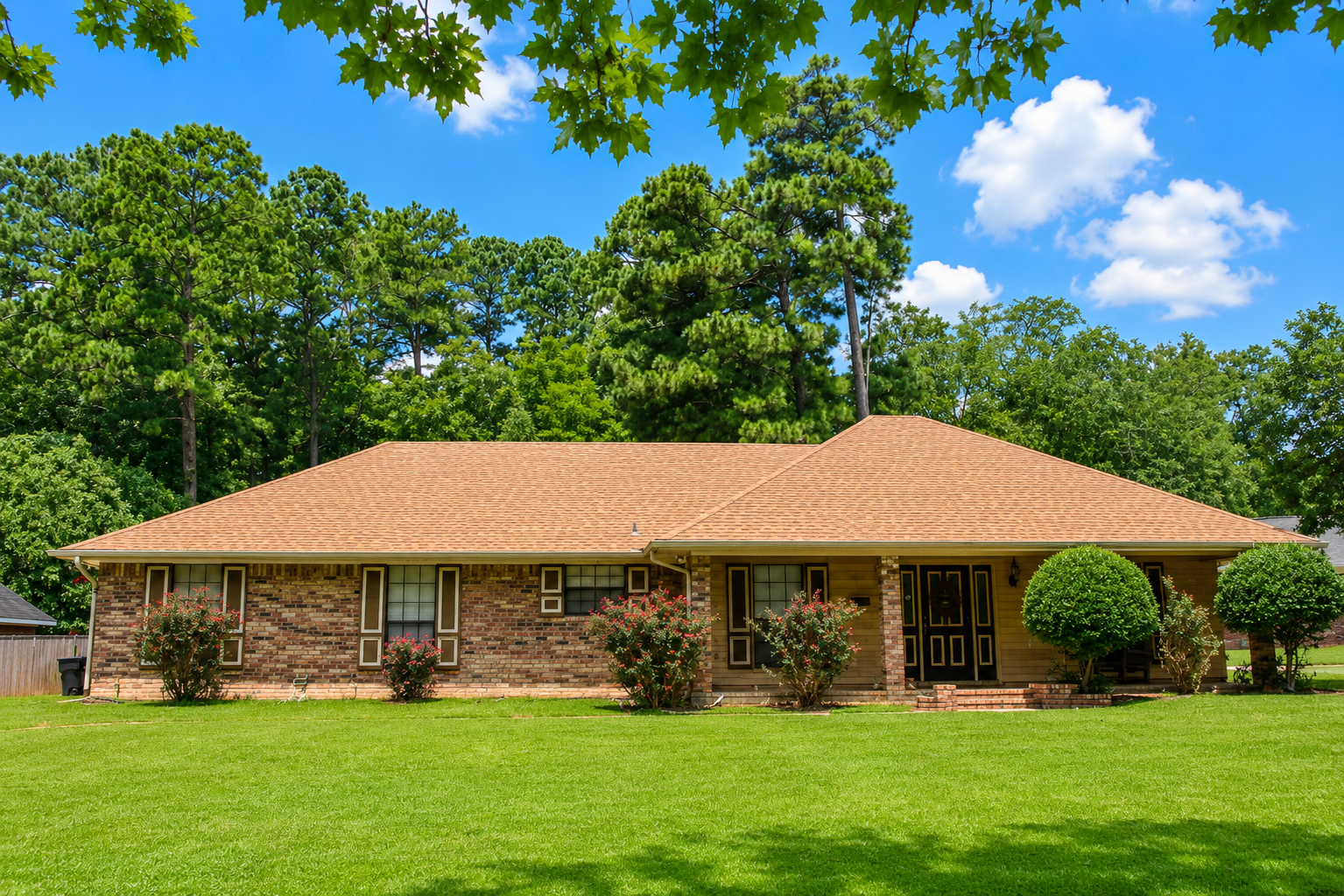 Single-story brick house with tan roof and green lawn under a blue sky, surrounded by trees