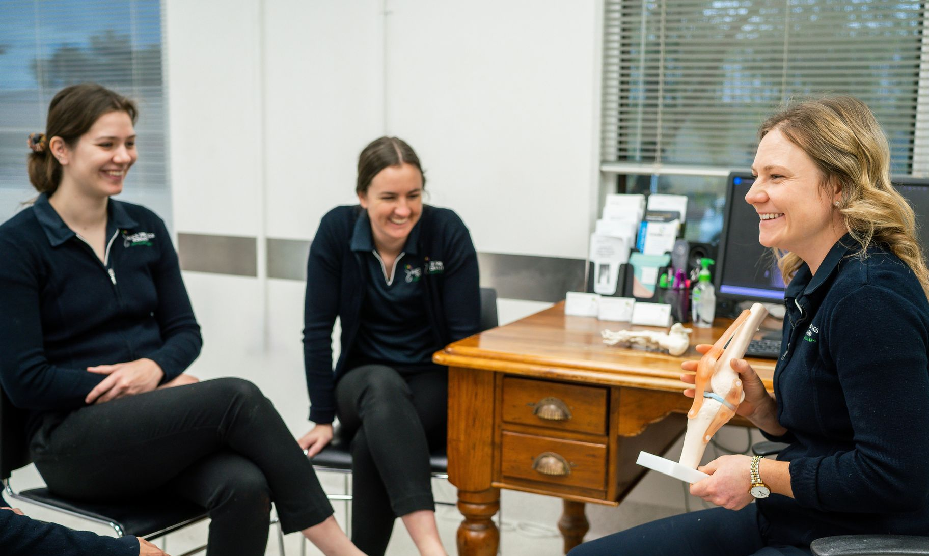 Three women in navy shirts laughing, one holding a knee model, seated around a small table in an office. — Alice Springs Podiatry in Ciccone, NT