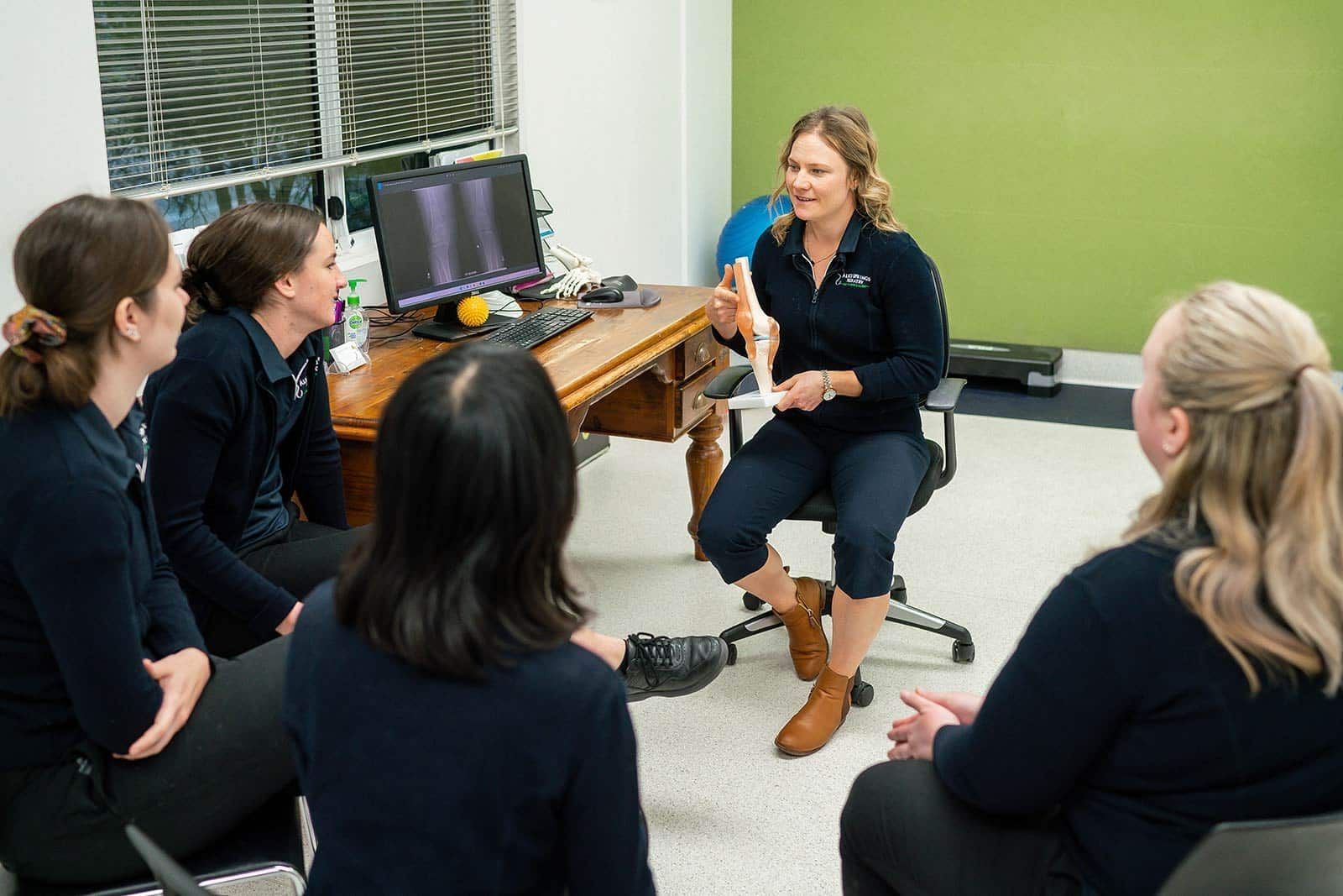 A woman presents a model to a group of four seated colleagues in a room; a treadmill is visible in the background. — Alice Springs Podiatry in Ciccone, NT