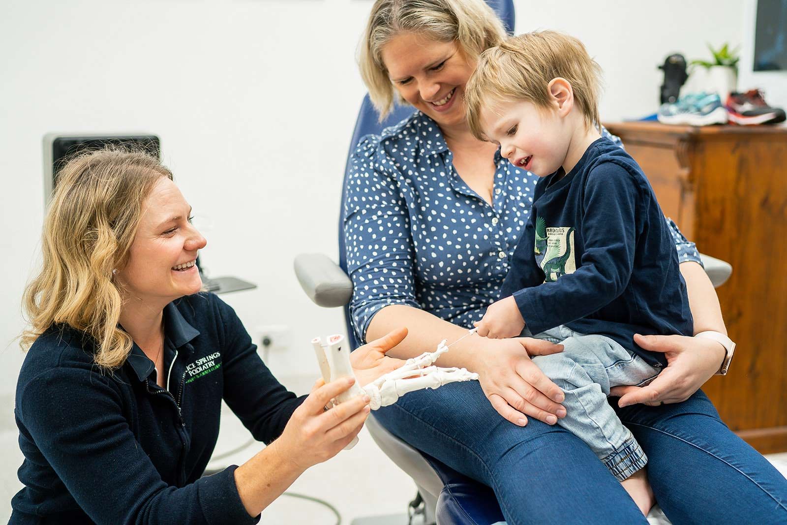 Woman shows a child a skeletal model, held by the child's mother. They are smiling in an office setting. — Alice Springs Podiatry in Ciccone, NT