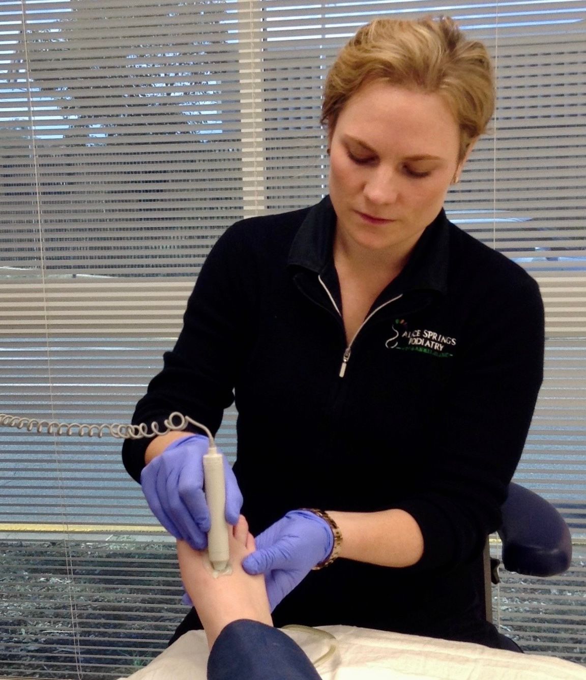 A woman wearing a black top and gloves uses a handheld device on a foot. Window blinds are in the background. — Alice Springs Podiatry in Ciccone, NT