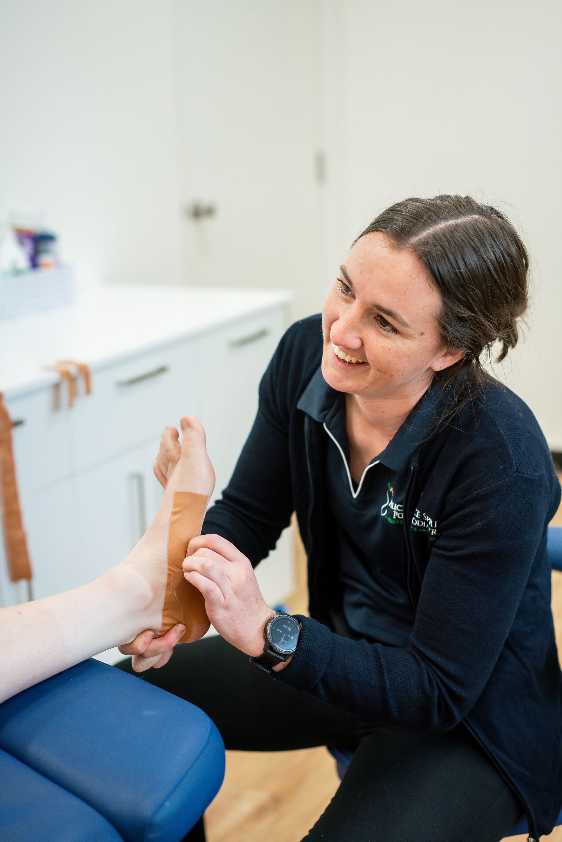 Woman in a clinic has her ankle assessed by a kneeling medical professional. The patient is seated. — Alice Springs Podiatry in Ciccone, NT