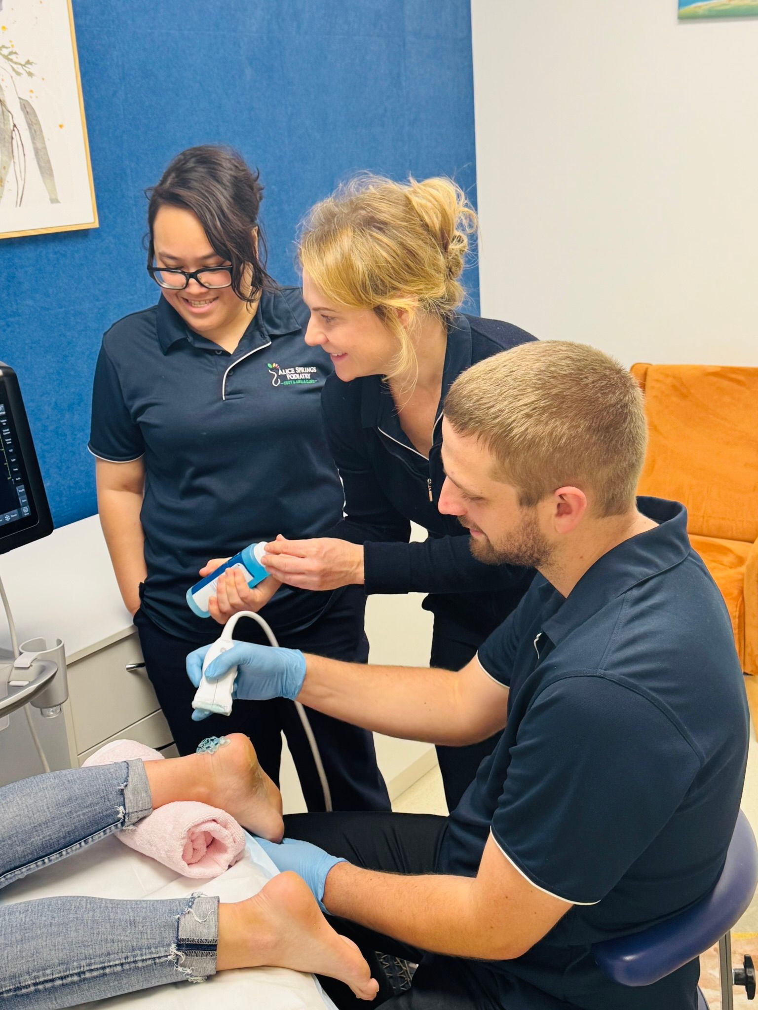 Three medical professionals examining a patient's foot with an ultrasound machine in a clinic. Two women and a man are smiling. — Alice Springs Podiatry in Ciccone, NT