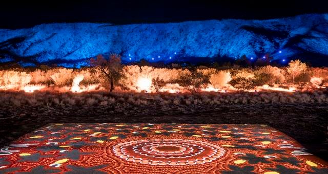 A night view of Uluru illuminated with blue and orange lights, with an Aboriginal artwork projected on the ground. — Alice Springs Podiatry in Ciccone, NT