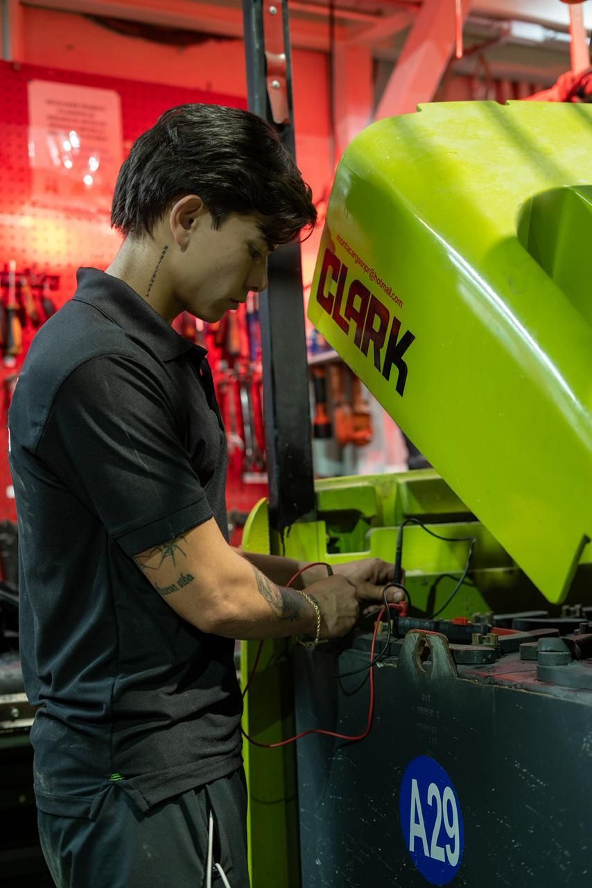Mecánico trabajando en una carretilla elevadora Clark verde lima, manipulando componentes eléctricos. Herramientas al fondo, en un entorno de taller.