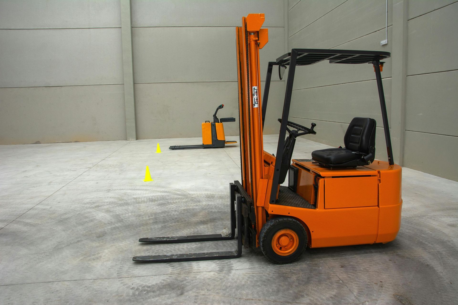 Carretilla elevadora naranja en un almacén con una pequeña transpaleta al fondo.