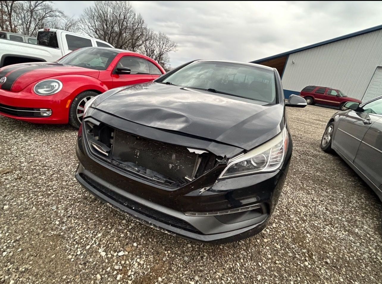 a black car with a damaged front end is parked in a gravel lot .