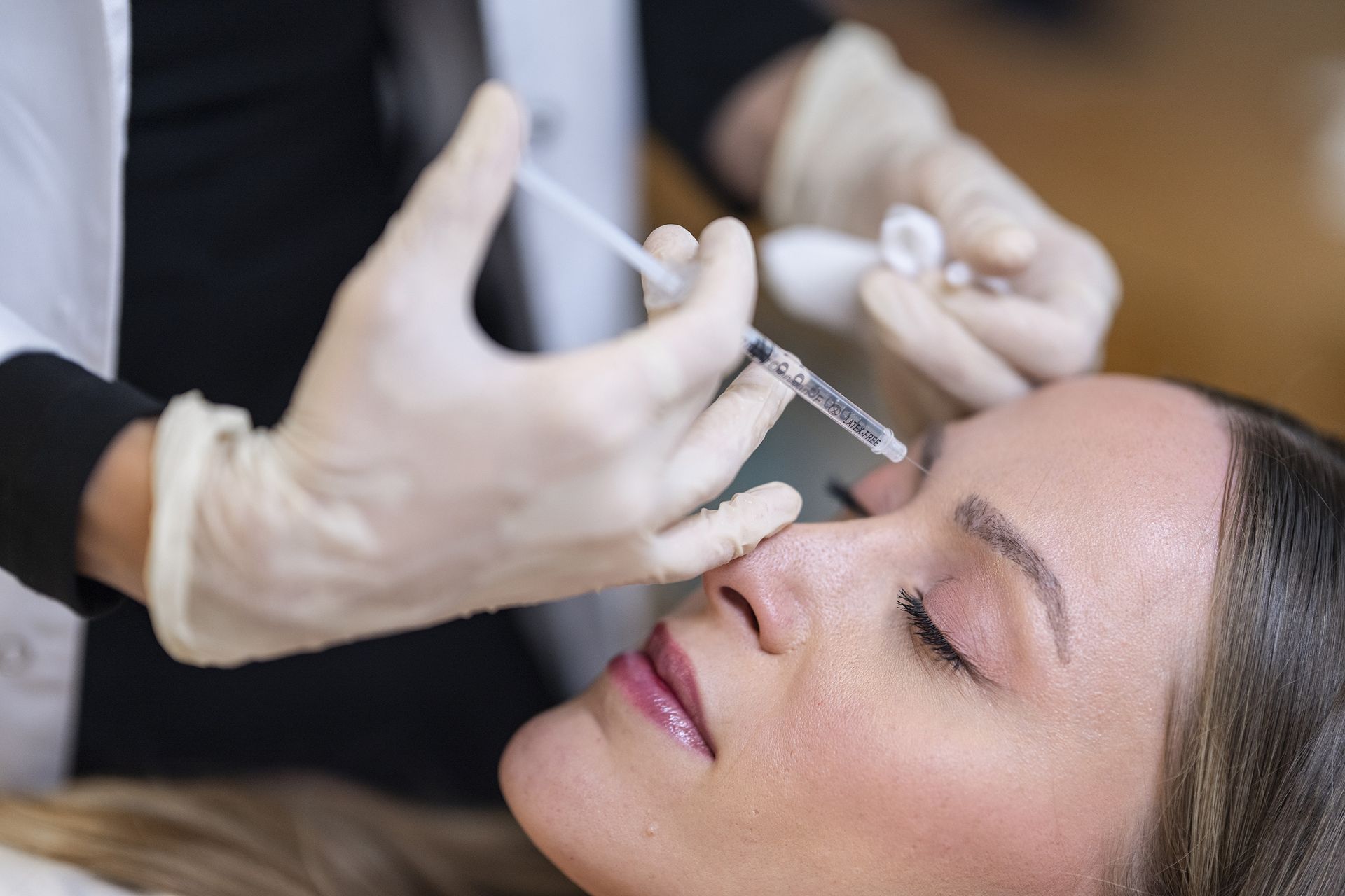 Hände mit Handschuhen, die einer Frau während einer kosmetischen Behandlung mit einer Spritze in die Stirn injizieren