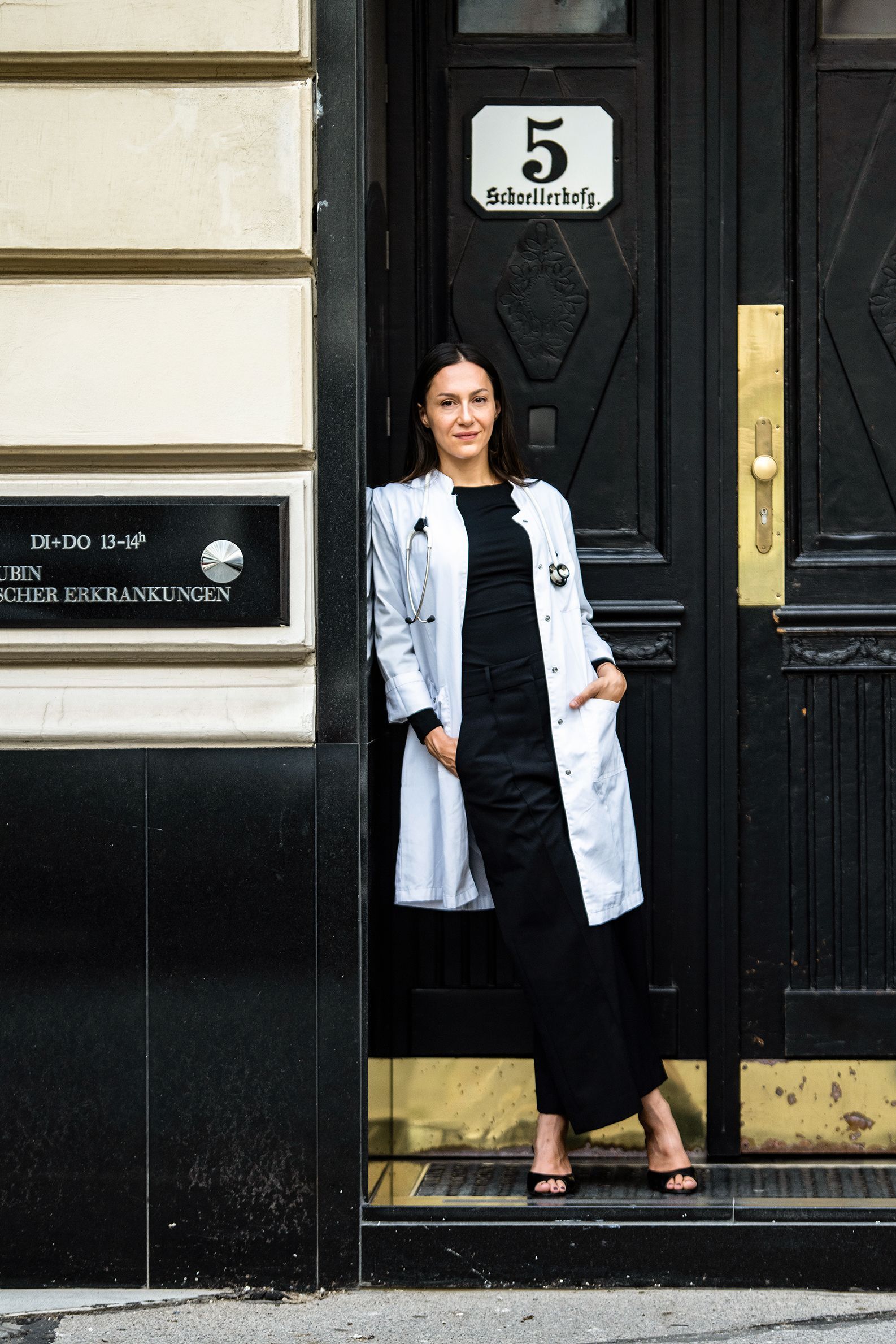 Person steht in einer Türöffnung und trägt einen langen weißen Mantel und ein schwarzes Outfit