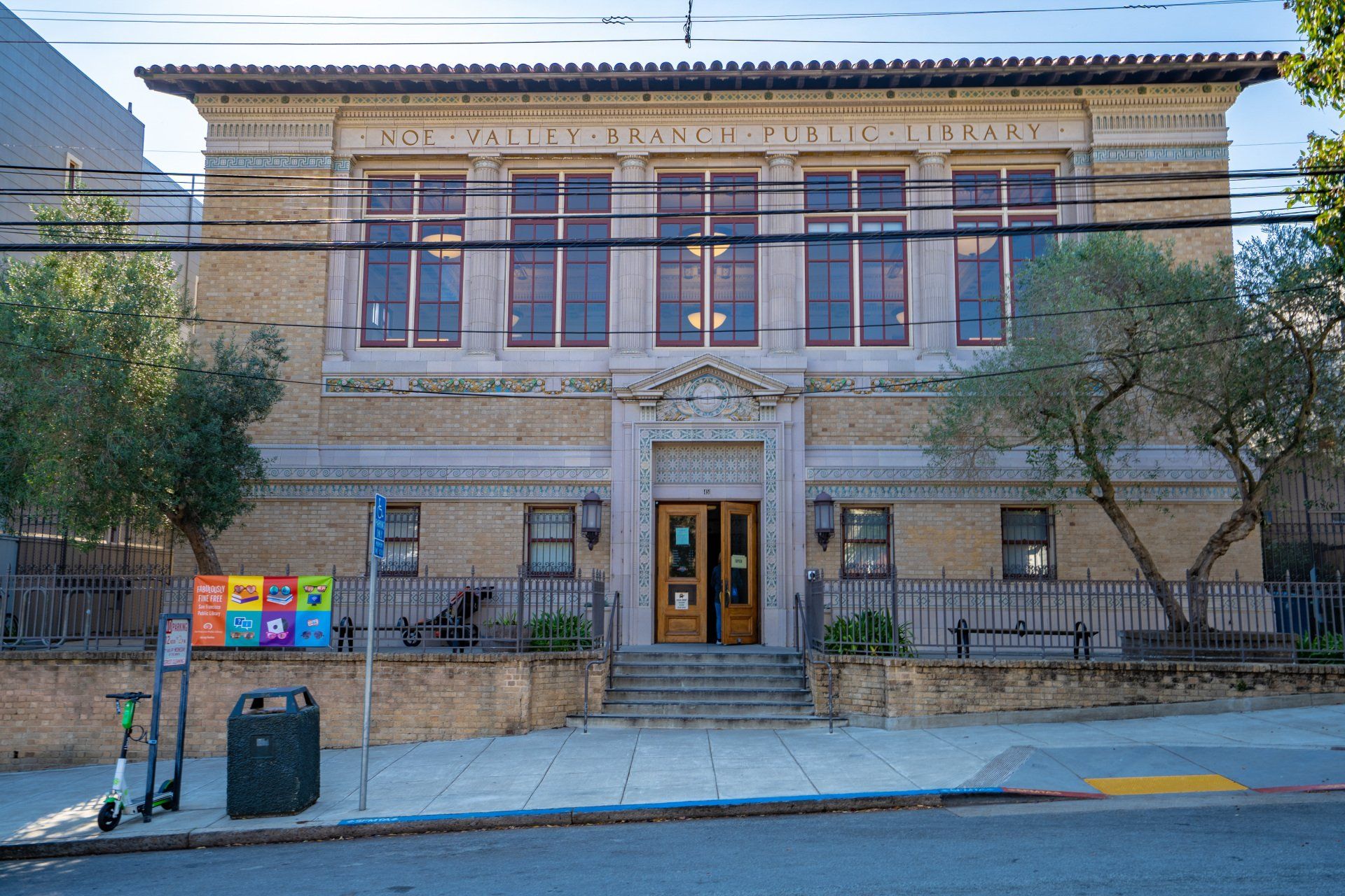 Exterior of a two-story library with a tan facade, steps leading to the entrance, and a sign reading
