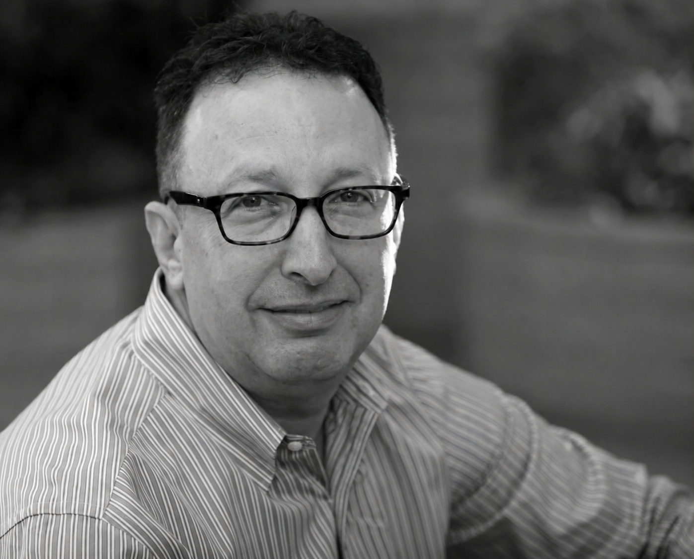 Man in glasses and shirt smiling outdoors against a blurred background